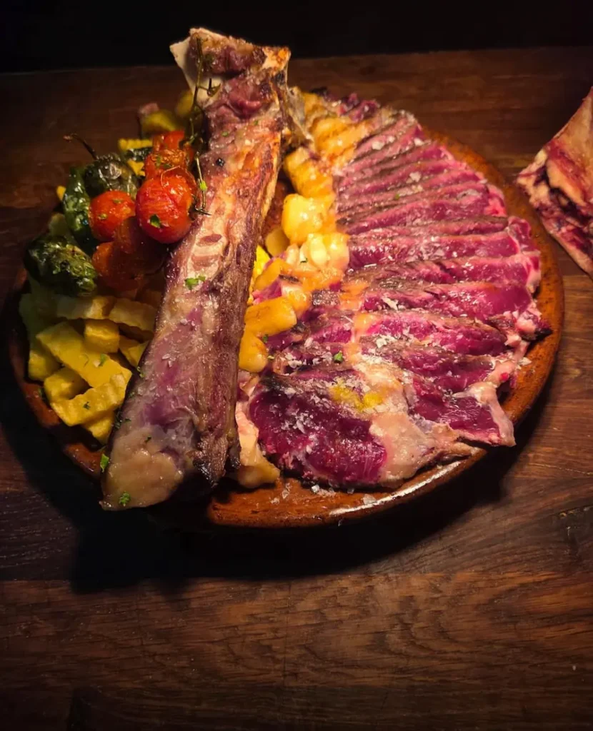 Chuletón Premium Red Meat cortado en láminas y servido al punto, acompañado de patatas fritas caseras y pimientos rojos asados sobre plato de barro.